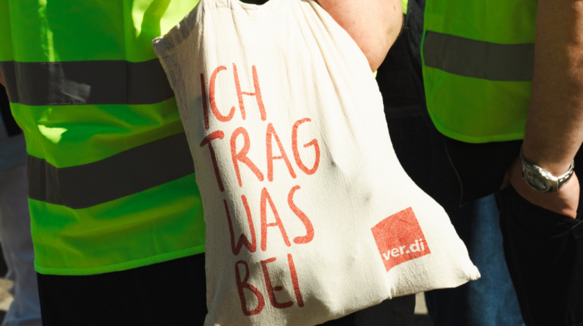 ver.di-Stoffbeutel, den jemand trägt, auf dem steht "Ich trag was bei"