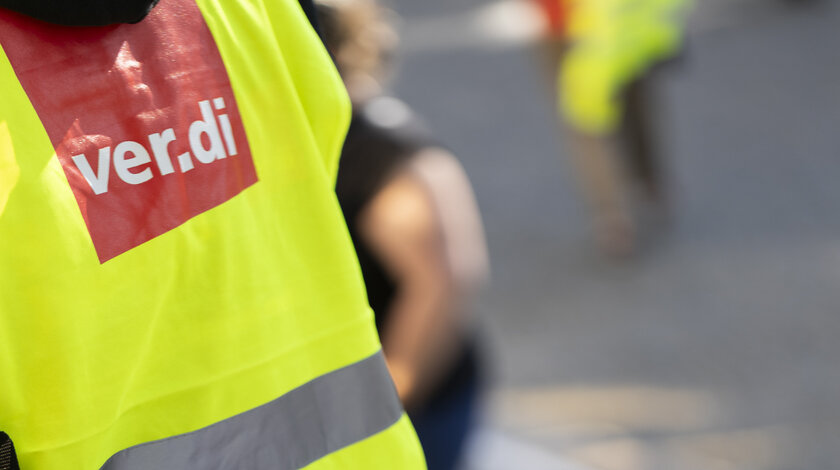 Eine Person von hinten, die eine gelbe Warnweste sowie ein Megafon trägt